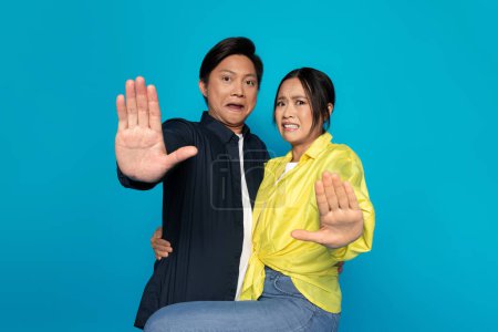 An Asian millennial couple looks shocked and defensive, holding up their hands in a stop gesture, portraying a need for personal space or rejection, against a blue background