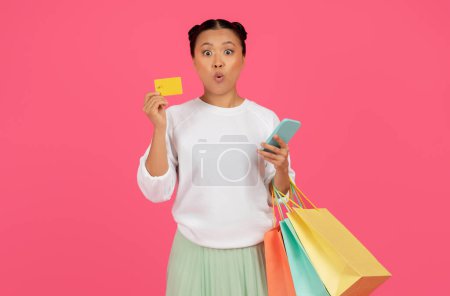 Photo for Shocked Asian Woman Holding Smartphone, Credit Card And Shopping Bags While Posing Over Pink Background, Surprised Female Enjoying Making Purchases In Internet, Emotionally Reacting To Online Sales - Royalty Free Image