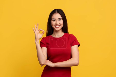 Foto de Positiva joven taiwanesa bastante joven mostrando gesto bien y sonriendo a la cámara, posando aislado en el fondo del estudio amarillo. Atractiva mujer asiática recomendando servicio o producto - Imagen libre de derechos