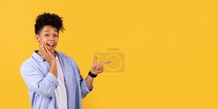 Téléchargez les photos : Étonné homme noir avec la main sur la joue pointant vers le côté à l'espace libre avec une expression joyeuse, sur fond jaune vif, suggérant idée fraîche ou découverte, panorama - en image libre de droit