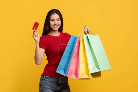 Happy attractive young chinese woman customer showing purchases colorful paper bags and red plastic credit card, smiling at camera, isolated on yellow background. Easy shopping, retail