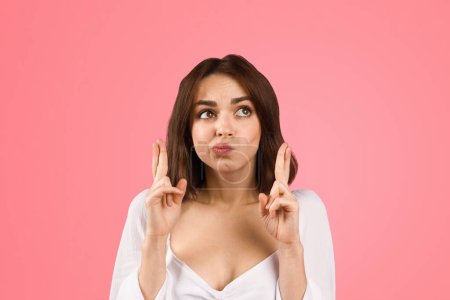 Photo for A young woman in a white top makes a hopeful gesture, crossing her fingers on both hands with a playful pout, expressing wishes or anticipation, against a pink background - Royalty Free Image