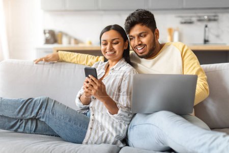 Photo for Engaged indian couple relaxing on couch with smartphone and laptop, happy young eastern spouses enjoying their leisure time at home, browsing new app or scrolling social networks, free space - Royalty Free Image