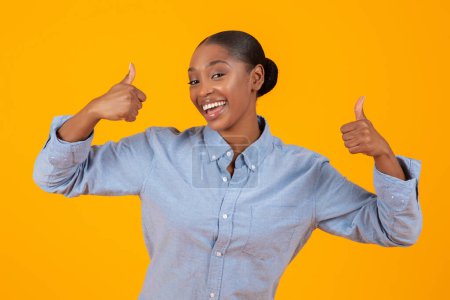 Téléchargez les photos : Ça me plaît. Portrait d'une femme noire joyeuse dans un geste décontracté pouce vers le haut Symbole, approuvant une grande offre, posant le port de chemise en denim debout sur fond jaune dans Studio - en image libre de droit