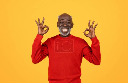 Fröhlicher älterer afrikanisch-amerikanischer Mann mit weißem Bart macht mit beiden Händen das OK-Zeichen, lächelt fröhlich, trägt einen roten Pullover vor schlichtem gelben Studiohintergrund