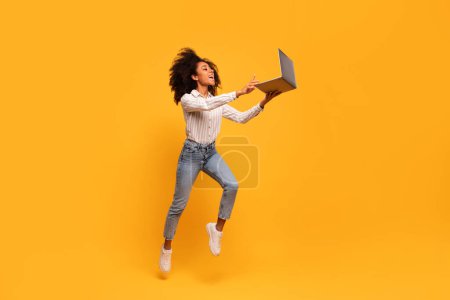 Vivacious young black woman with curly hair in mid-jump, holding laptop outstretched with look of triumph, set against bright yellow background