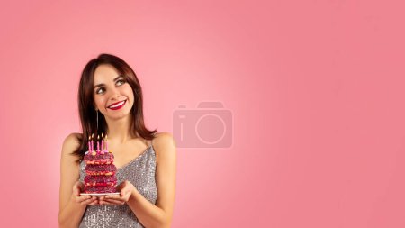 Photo for A smiling woman with brown hair and red lipstick, wearing a glittery silver dress, is holding a colorful donut stack with lit candles against a soft pink background - Royalty Free Image