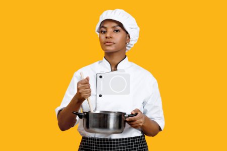 Photo for Serious black female chef holding wooden spoon and stainless steel pot, confident african american woman cook ready for culinary action, standing against monochrome yellow background, copy space - Royalty Free Image