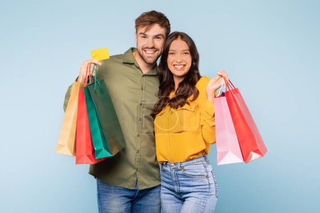 Pareja radiante mostrando éxito de compras con bolsas de colores y tarjeta de crédito, lo que sugiere una experiencia de venta satisfactoria en fondo azul