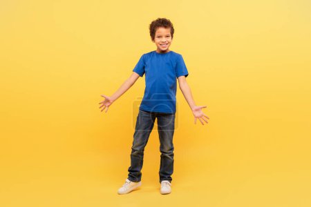 Photo for Smiling young boy with curly hair in blue t-shirt with arms wide open, expressing happiness, standing against bright yellow background - Royalty Free Image