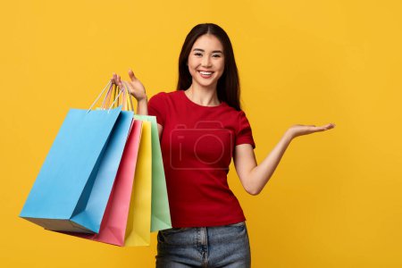 Consumerism, retail. Smiling asian woman with colorful shopping bags showing something invisible product, good on her hand palm, copy space, isolated on yellow studio background