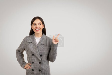 Foto de Mujer joven entusiasta en blazer a cuadros apunta alegremente a un lado en el espacio libre, mostrando algo emocionante, con una gran sonrisa contra el fondo gris - Imagen libre de derechos