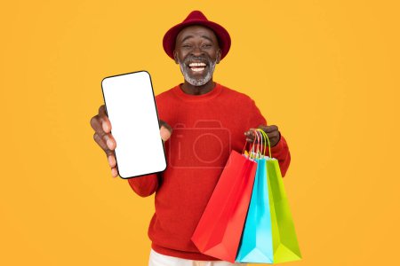 Téléchargez les photos : Joyeux homme noir âgé en pull rouge et chapeau, montrer smartphone écran vierge et tenir des sacs à provisions colorés, représentant achats en ligne ou l'excitation de vente, sur fond jaune vif - en image libre de droit