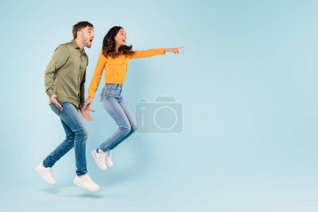 Téléchargez les photos : Jeune couple énergique à mi-saut pointant vers l'espace libre avec excitation, affichant un sentiment de plaisir et de découverte sur fond bleu vif, bannière - en image libre de droit