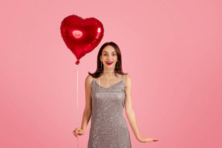 Téléchargez les photos : Femme caucasienne heureuse surprise et ravie dans une robe à paillettes argentée glamour tenant un ballon rouge en forme de cœur, avec un geste ouvert sur un fond rose, studio, annonce et offre - en image libre de droit