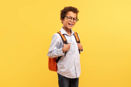 Foto de Feliz colegial negro con el pelo rizado, con gafas y sonriendo a la cámara, llevando una mochila naranja sobre un fondo amarillo soleado - Imagen libre de derechos