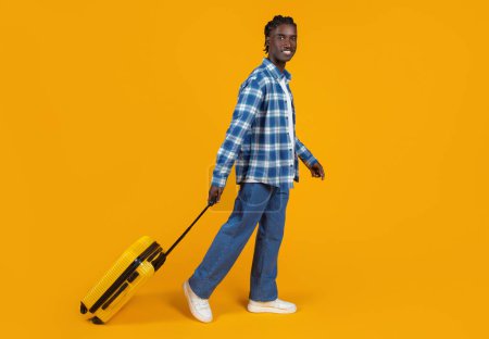 Photo for Smiling young black man casually walking with yellow suitcase, happy guy dressed in plaid shirt and jeans, looking ready for adventures, posing against bright orange background, enjoying travels - Royalty Free Image