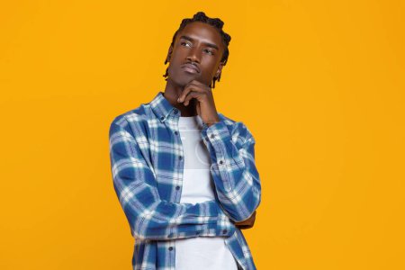 Portrait of thoughtful young black man touching chin and looking away, african american guy in checkered shirt looking contemplative, standing isolated against yellow studio background, copy space