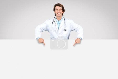 Téléchargez les photos : Médecin masculin joyeux avec stéthoscope pointant vers le bas à grande bannière vierge, parfait pour les messages de santé, annonces ou publicités, espace libre - en image libre de droit