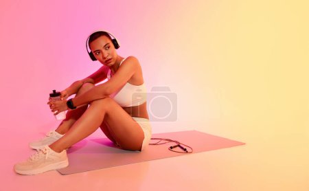 Foto de Mujer latina milenaria enfocada con un corte de zumbido sentada en una esterilla de yoga, sosteniendo una botella de agua, con auriculares y una cuerda de salto cerca, sobre un fondo rosa y amarillo degradado - Imagen libre de derechos
