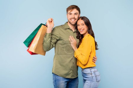 Téléchargez les photos : Homme et femme souriants tenant des sacs colorés, regardant satisfaits de leurs achats, partageant un moment de vente au détail joyeux sur fond bleu clair - en image libre de droit