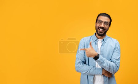 Foto de Un hombre carismático con barba y gafas sonríe y señala de lado con su mano izquierda, dando una impresión de amabilidad y accesibilidad contra un fondo naranja - Imagen libre de derechos