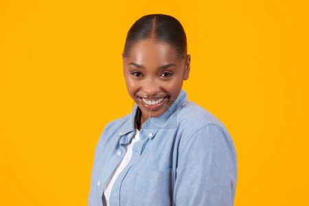 Téléchargez les photos : Portrait de joyeux jolie dame afro-américaine en chemise en denim souriant avec confiance à la caméra sur fond de studio jaune, exprimant des émotions positives et la publicité grande offre - en image libre de droit