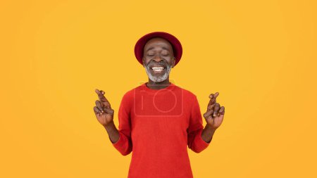 Photo for Content senior black man with closed eyes and a serene smile, wearing a red sweater and hat, crossing his fingers for good luck, expressing hope and positivity on a vibrant yellow background - Royalty Free Image