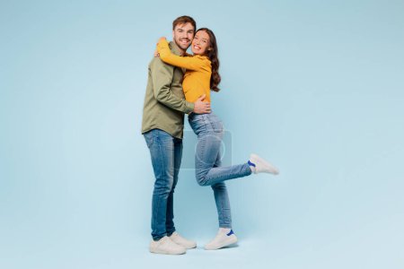 Téléchargez les photos : Couple aimant dans une étreinte heureuse, avec l'homme soulevant la femme de ses pieds, les deux souriant joyeusement, montrant l'affection et le bonheur sur fond bleu apaisant - en image libre de droit