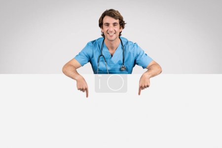 Téléchargez les photos : Infirmière souriante en blouse bleue pointant vers une bannière blanche vide, idéale pour le placement de texte ou de graphiques, sur un fond gris clair - en image libre de droit