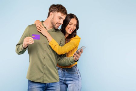 Foto de Pareja sonriente dedicada a las compras en línea, con el hombre sosteniendo la tarjeta de crédito y la navegación en el teléfono inteligente, cónyuges disfrutando del comercio electrónico en el fondo azul - Imagen libre de derechos