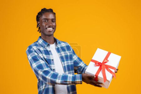 Happy young black man extending white gift box with red ribbon, cheerful african american guy offering present with friendly smile, standing against bright yellow background, free space