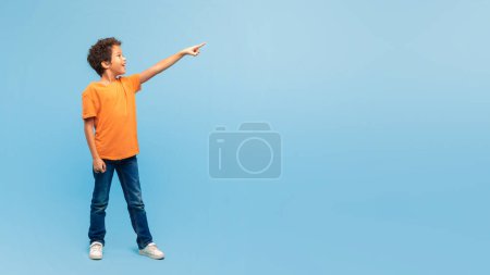Téléchargez les photos : Joyeux garçon aux cheveux bouclés, portant une chemise orange et un jean, pointant vers le haut vers l'espace libre avec une expression joyeuse, sur un fond bleu serein, panorama - en image libre de droit