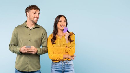 Téléchargez les photos : Homme souriant tenant smartphone et femme avec carte de crédit, effectuant éventuellement une transaction en ligne ou planifiant un achat, regardant l'espace libre sur fond bleu - en image libre de droit