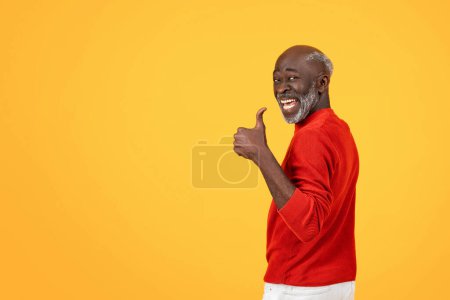 Téléchargez les photos : Enthousiaste homme noir senior avec une barbe blanche donnant un signe pouces vers le haut, se retournant avec un grand sourire, dans un pull rouge sur un fond jaune uniforme, montrant l'approbation et la satisfaction - en image libre de droit