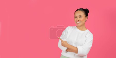 Foto de Mujer asiática enérgica haciendo un gesto a la derecha en el espacio vacío, alegre coreana mujer demostrando lugar para su anuncio, de pie aislado sobre fondo rosa brillante, panorama - Imagen libre de derechos
