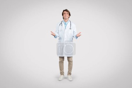Happy young male doctor in a lab coat with stethoscope presenting an open-handed welcoming gesture, standing against a plain gray background, full length