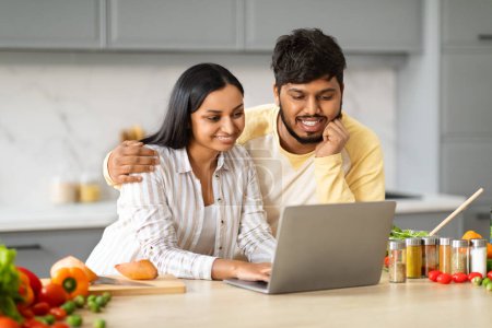 Photo for Diet, nutrition. Loving millennial indian couple vegetarians cooking together, checking healthy recipes on Internet. Eastern man and woman preparing delicious dinner, looking at computer screen - Royalty Free Image