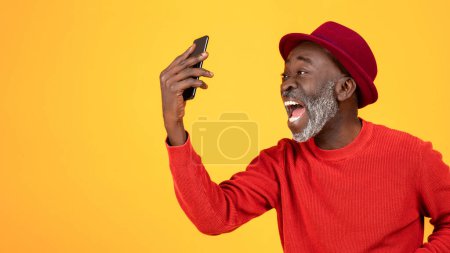 Photo for Happy or angry excited senior african american man in hat and red clothes, with open mouth, scream on phone, isolated on orange studio background, close up, panorama. Win, good news, quarrel - Royalty Free Image