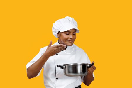 Photo for Satisfied black female chef smelling the flavor from a cooking pot, reflecting the taste success, happy african american cook woman standing against yellow background, indicating culinary delight - Royalty Free Image