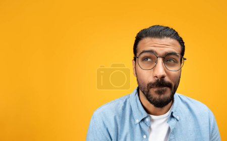 Pensive indian millennial man in glasses, look at free space, think, isolated on orange background, studio, close up. Create idea, ad and offer, sale and brainstorm, people emotions