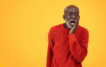 Téléchargez les photos : Homme noir âgé avec une barbe blanche touchant sa joue dans la douleur, peut-être avoir un mal de dents, avec une expression douloureuse, vêtu d'un pull rouge sur un fond jaune - en image libre de droit