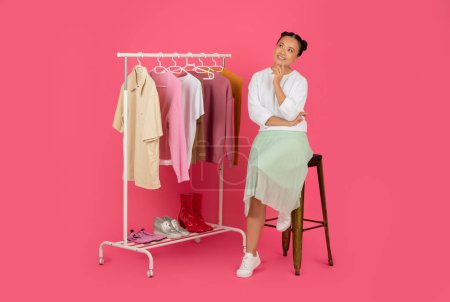 Photo for Fashion Idea. Pensive Smiling Asian Woman Sitting Near Clothing Rack, Thoughtful Korean Female Touching Chin And Looking Up, Thinking About Fashion And Shopping, Posing On Pink Studio Background - Royalty Free Image