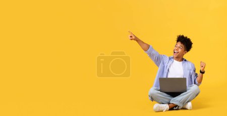 Photo for Excited male student in casual attire sitting cross-legged on yellow background, pointing upwards at free space and using laptop, showing look of inspiration, panorama - Royalty Free Image