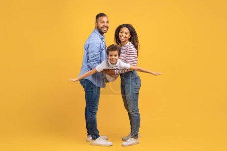Photo for Family Fun. Cheerful Black Mom And Dad Playing With Their Preteen Son, Bonding Together Over Yellow Studio Background, Happy African American Boy Pretending Flying, Spreading Hands, Imitating Plane - Royalty Free Image