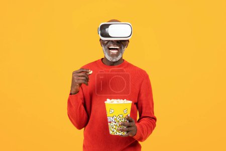 Téléchargez les photos : Souriant inspiré homme afro-américain senior en vr 3d lunettes, manger du pop-corn, regarder le film, profiter de collation et de temps libre, sur fond orange. Divertissement, nourriture, divertissement et cinéma - en image libre de droit