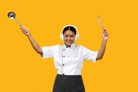 Foto de Chef negro sonriente bailando con utensilios de cocina en las manos, dirigiendo una orquesta, cocinera afroamericana feliz usando auriculares, escuchando música y divirtiéndose sobre fondo amarillo - Imagen libre de derechos