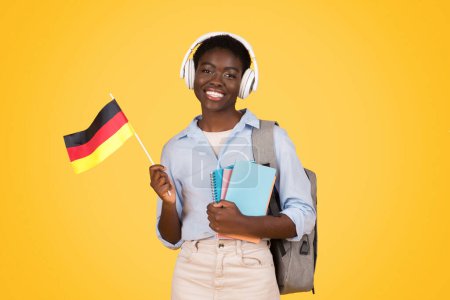 Photo for Happy teen african american woman student in wireless headphones, with books, flag of Germany, listen lesson, isolated on yellow studio background. Study and education exchange, learn German - Royalty Free Image