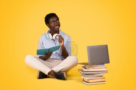 Photo for Smiling pensive teen black lady student sit with laptop on floor, read book, look at free space, isolated on yellow background, studio. Academic project, study online, education, create idea - Royalty Free Image