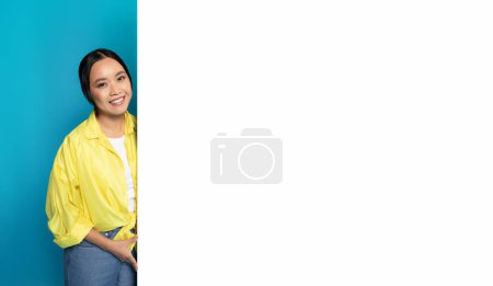 Foto de Mujer asiática sonriente con una camisa amarilla apoyada en una pancarta vertical blanca en blanco, mirando a la cámara con un comportamiento amistoso, sobre un fondo turquesa contrastante - Imagen libre de derechos